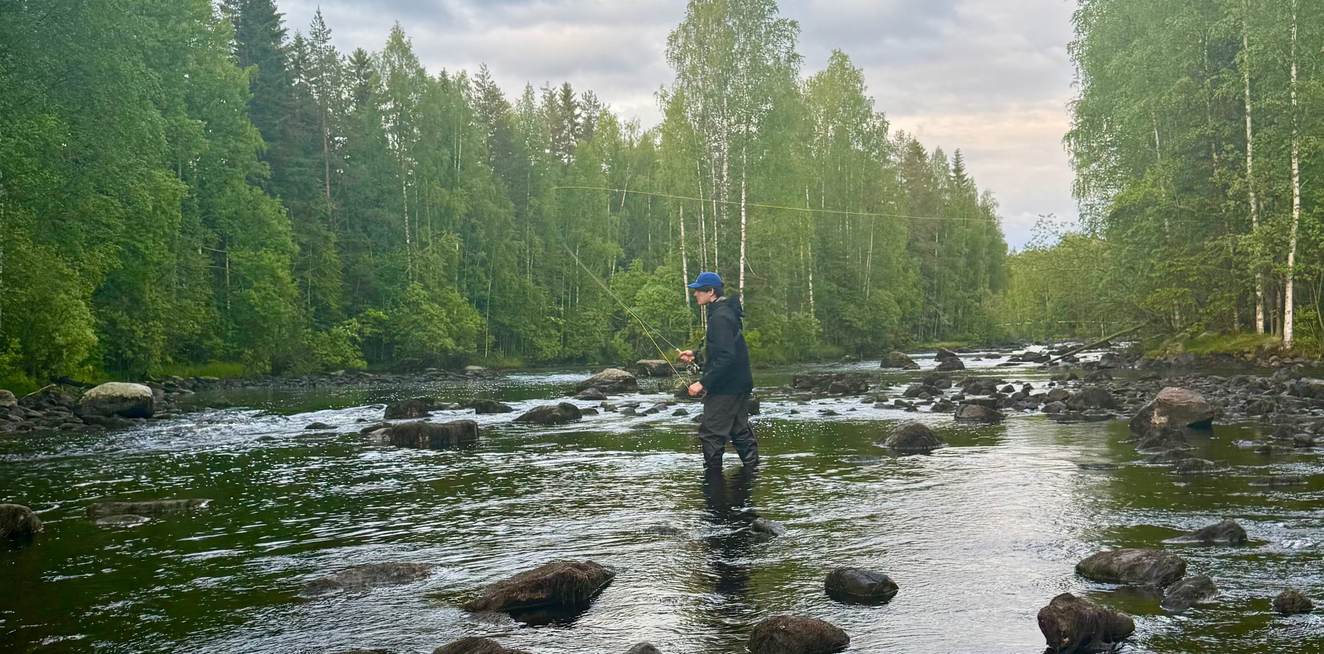 Person fishing in a river surrounded by trees on a cloudy day