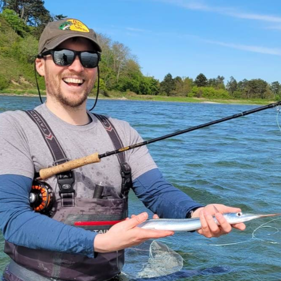 Man holding a garfish caught while fishing on the coast with trees in the background