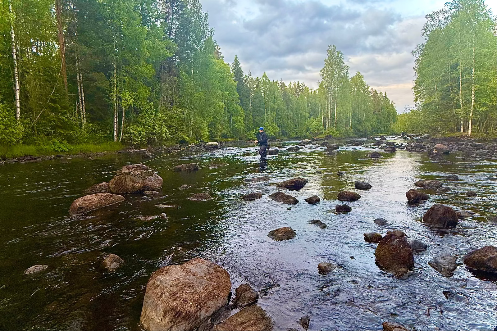 Person fishing in a river surrounded by trees on a cloudy day