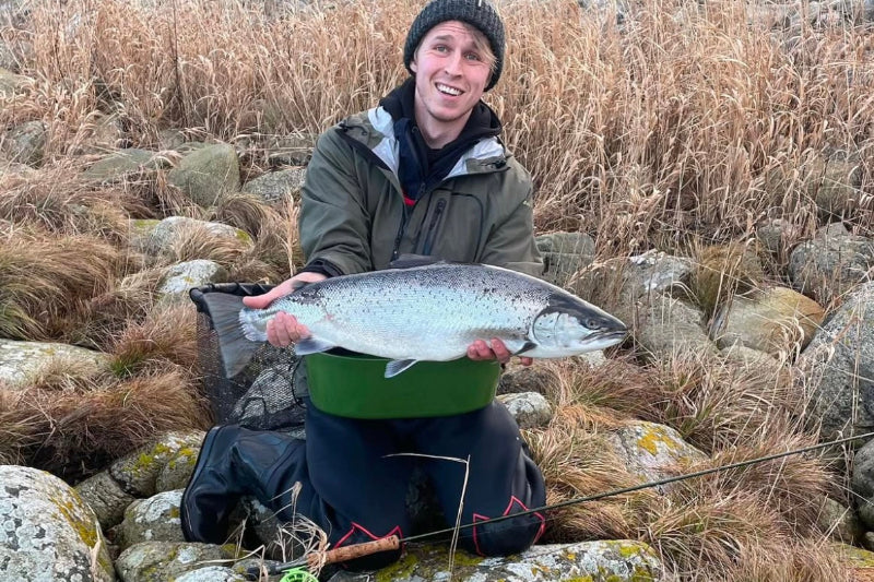 Person holding a large trout outdoors with dry grass and rocks in the background