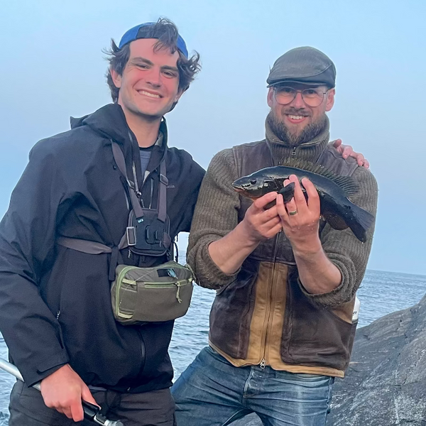 Two men standing outdoors by a body of water, one holding a fish.